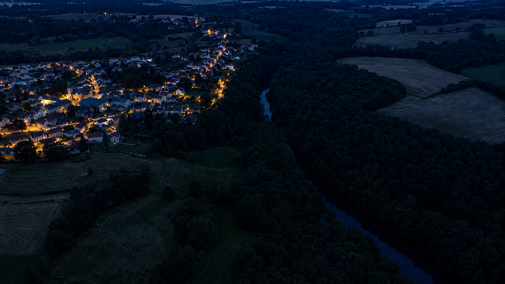 Vue nocturne d’une commune bordée par un corridor boisé et un cours d’eau laissés dans l’obscurité